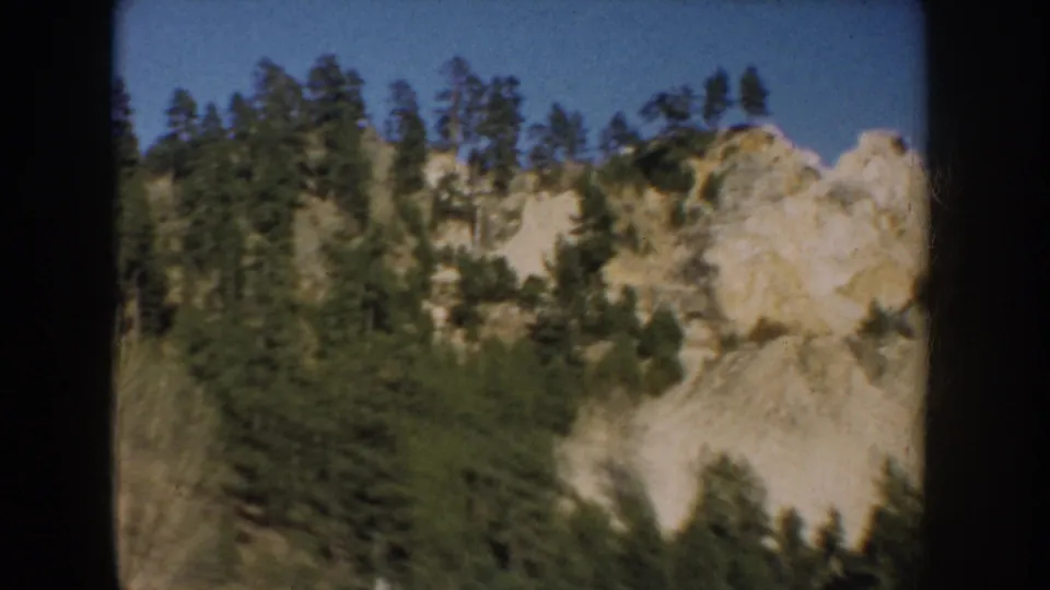 1953 South Dakota 8mm frame showing a pine-covered rocky slope with pale cliff sections below blue sky.