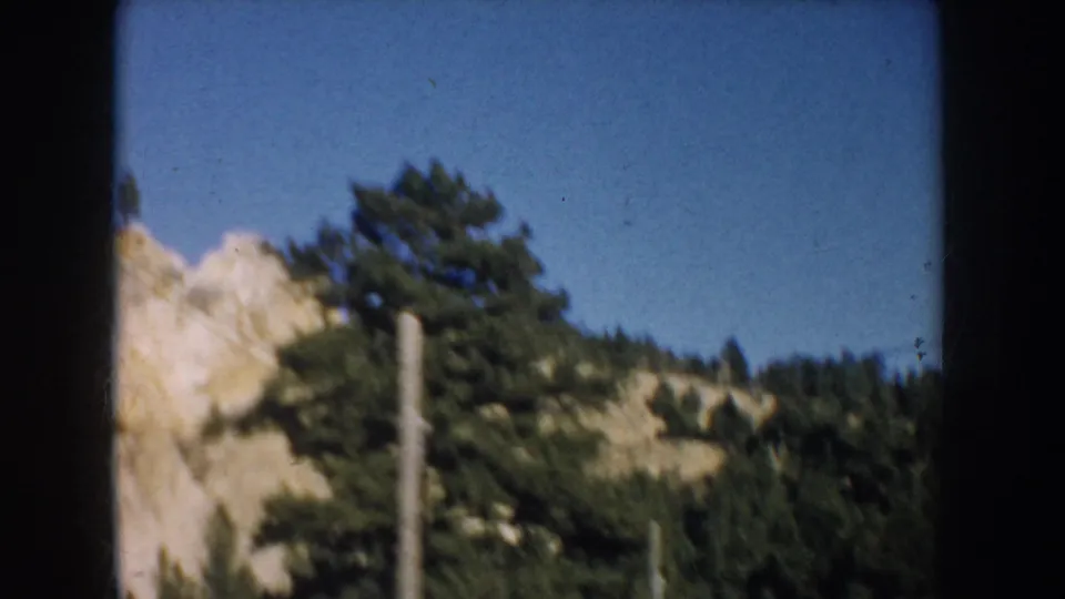 1953 South Dakota travel frame with evergreen trees and pale rock slope partly blocked by a dark vertical edge.