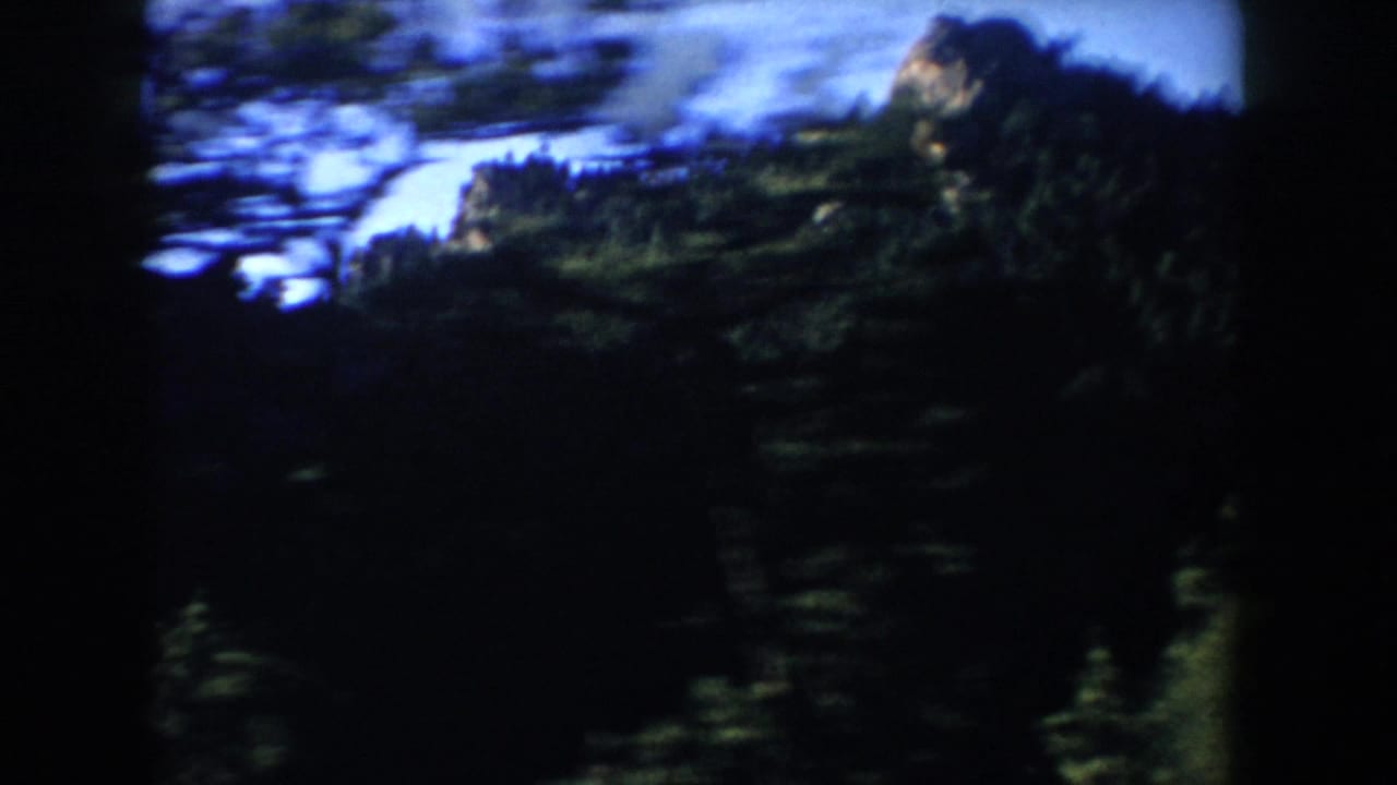 A moving home-movie frame from South Dakota shows dark pine forest in the foreground with rocky mountain shapes beyond.