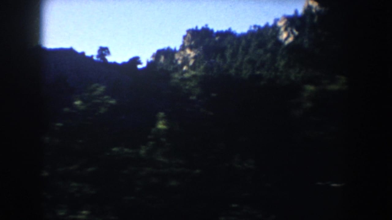 1953 South Dakota 8mm frame showing a shadowed forested hillside, tree silhouettes, and a bright blue sky.