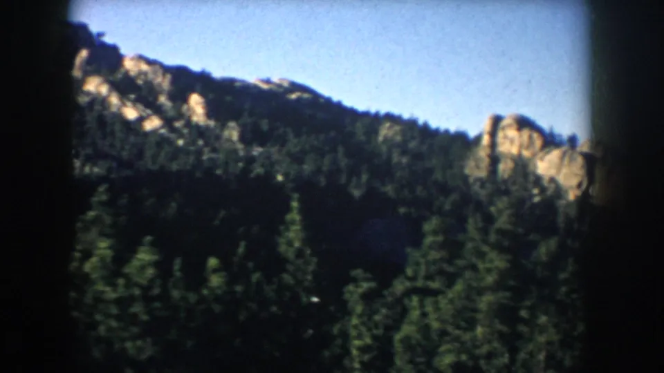 1953 South Dakota 8mm view of evergreen-covered slopes and pale rock formations beneath a clear blue sky.