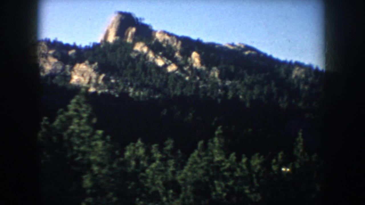 1953 South Dakota 8mm frame showing rugged rock formations above dense evergreens from a passing travel perspective.
