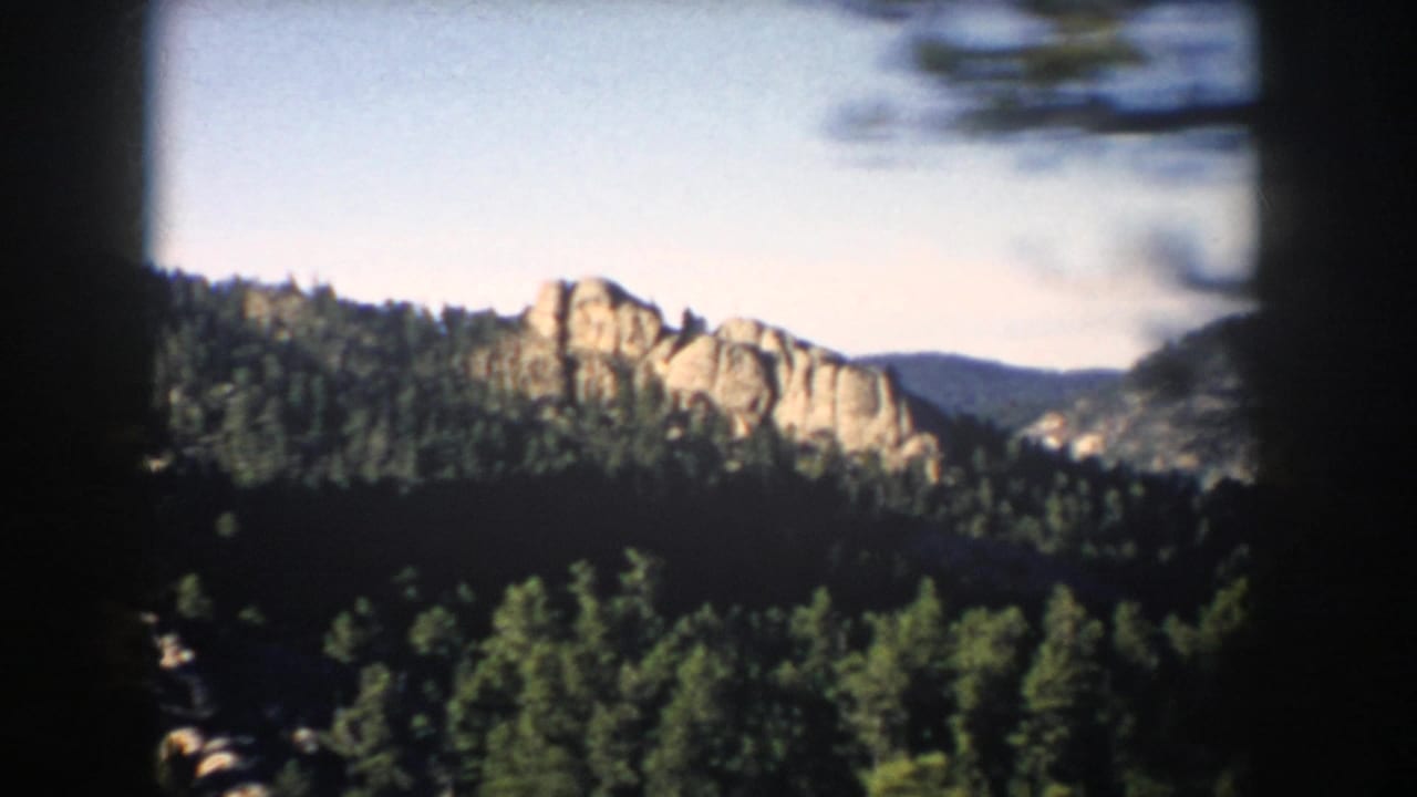 1953 South Dakota landscape frame showing pale rock formations rising above dark green pine forest beneath clear sky.