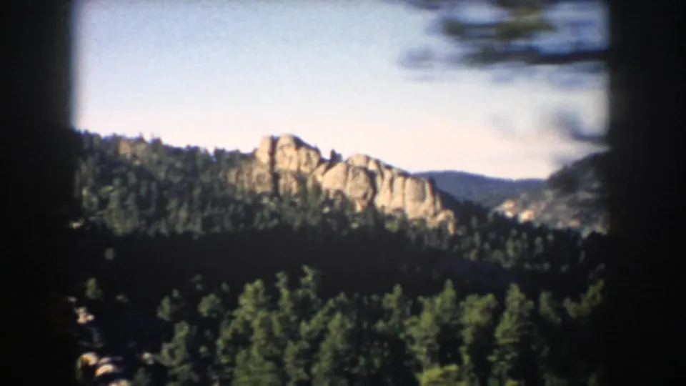 1953 South Dakota landscape frame showing pale rock formations rising above dark green pine forest beneath clear sky.