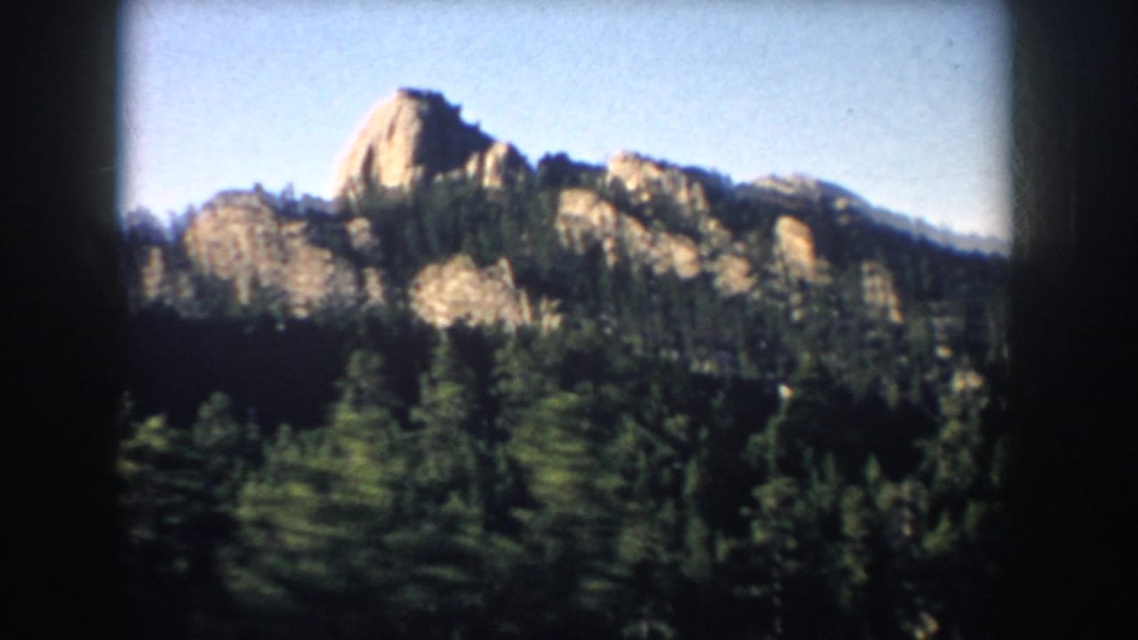 1953 South Dakota home-movie view of a prominent sunlit rock formation rising above pine forest, with soft 8mm color and edge darkening.