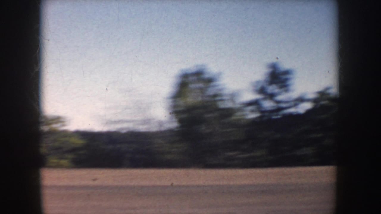 1953 South Dakota road-trip view with blurred fields, trees, and dark vehicle framing from a moving car.