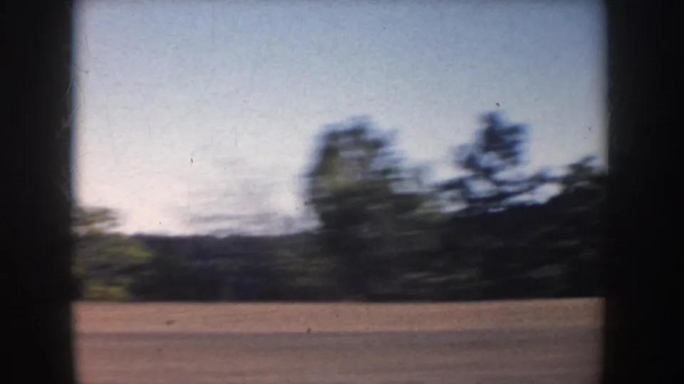 1953 South Dakota road-trip view with blurred fields, trees, and dark vehicle framing from a moving car.