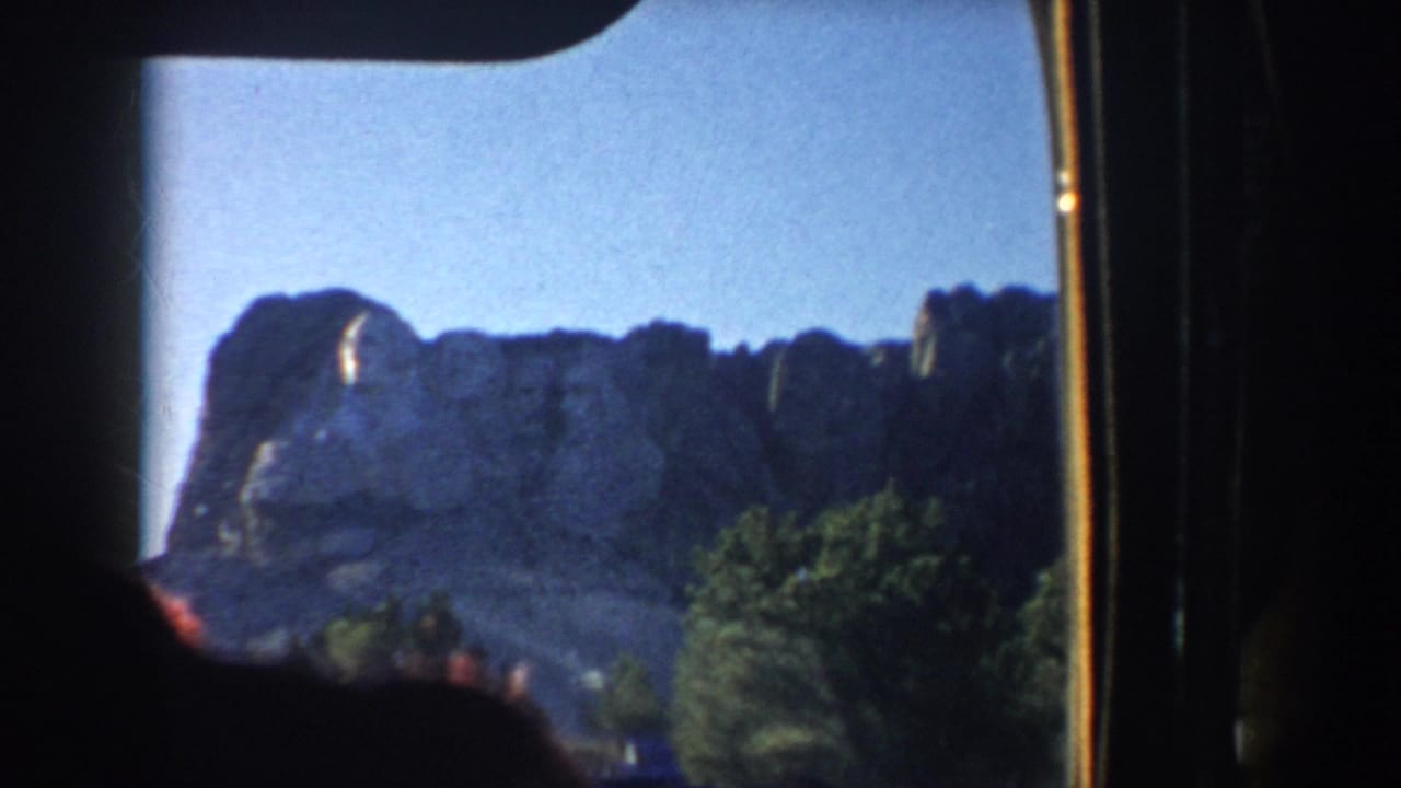 Mount Rushmore granite face appears against blue sky, partly framed by trees and the edge of the vehicle interior.
