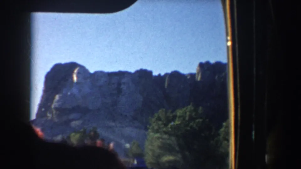 Mount Rushmore granite face appears against blue sky, partly framed by trees and the edge of the vehicle interior.