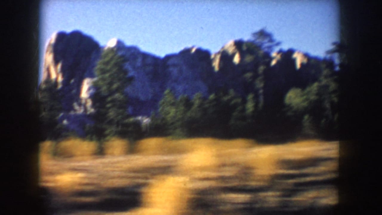 Mount Rushmore is seen across dry roadside grass, with granite faces, evergreen trees, and blue sky in 1953 home-movie footage.