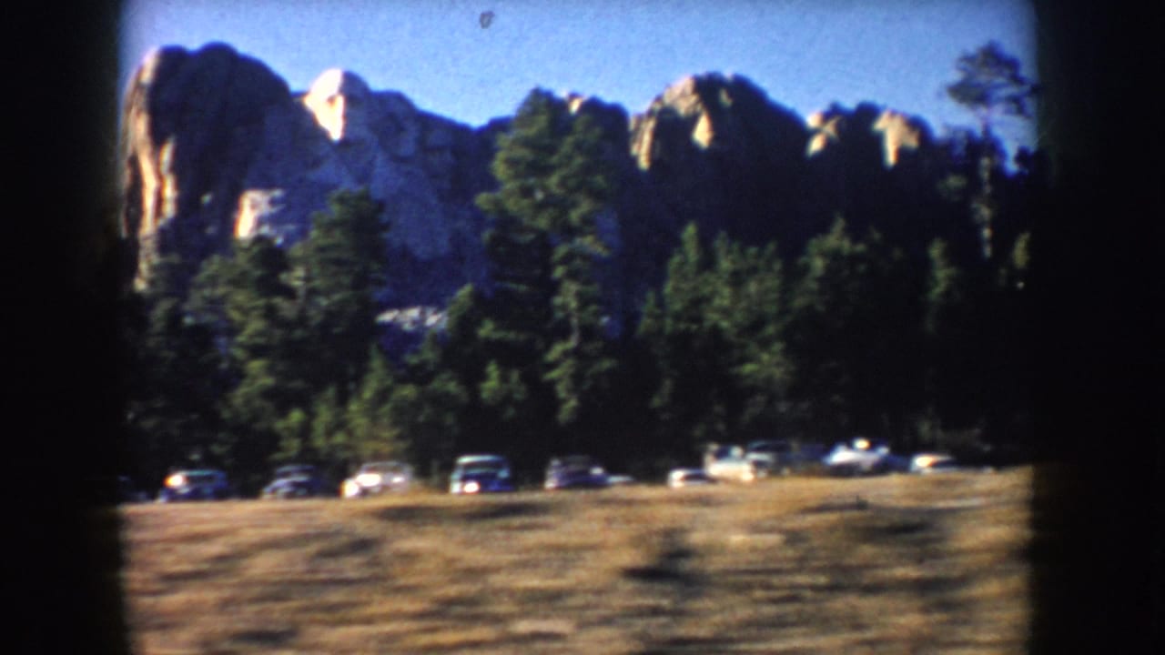 1953 South Dakota home-movie frame showing Mount Rushmore beyond pine trees and a row of period cars near the road.