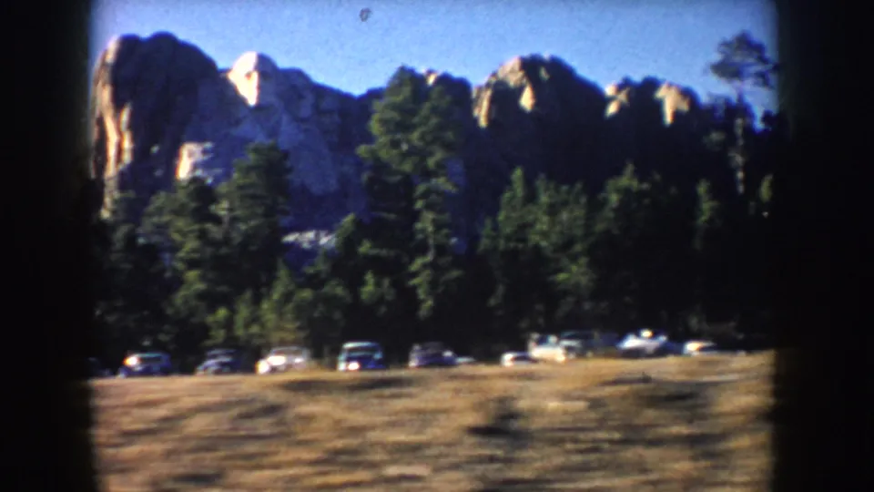 1953 South Dakota home-movie frame showing Mount Rushmore beyond pine trees and a row of period cars near the road.