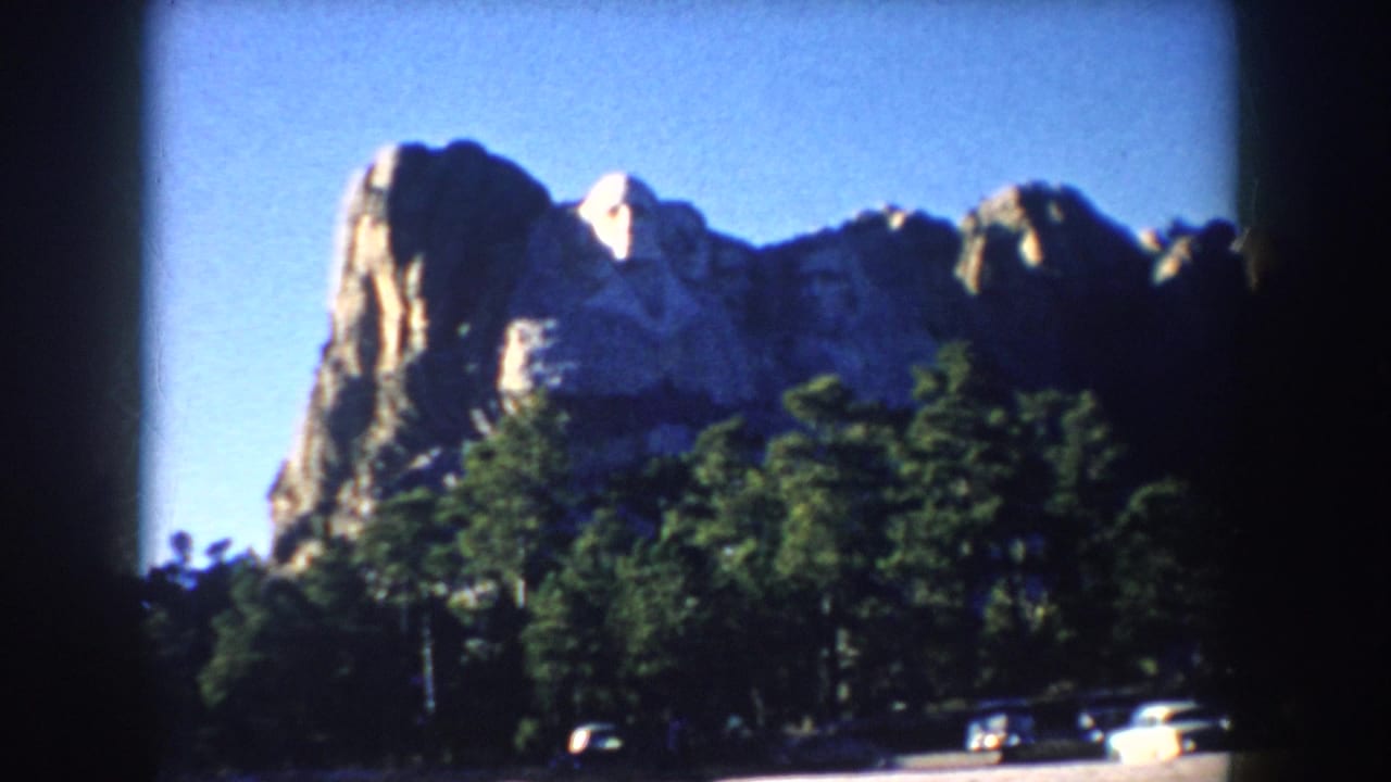 Closer 1953 home-movie view of Mount Rushmore rising above pine trees, with a few roadside cars visible below.