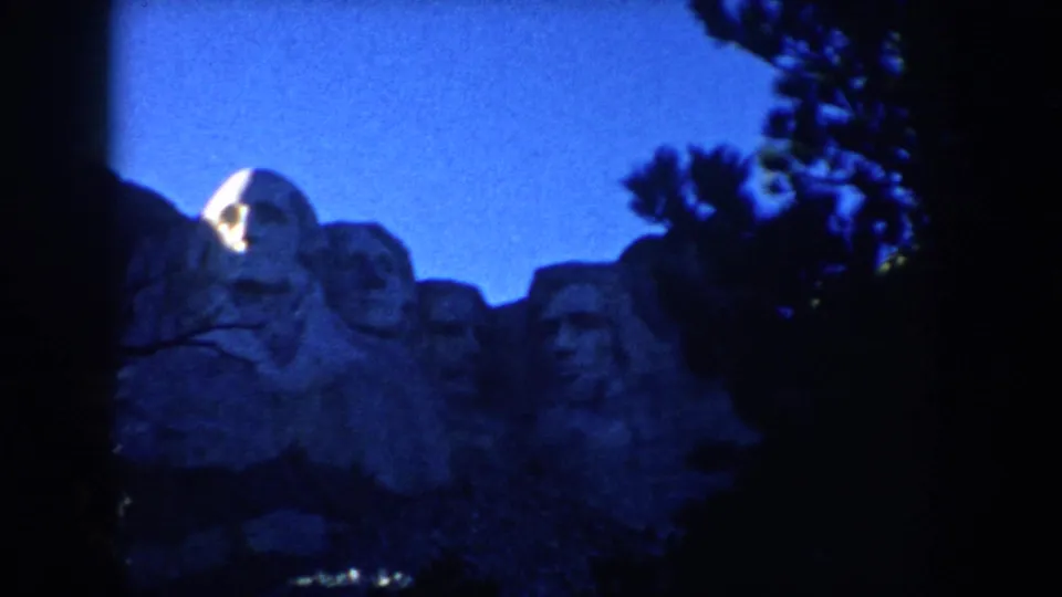 1953 South Dakota home-movie view of Mount Rushmore at night, with illuminated carved faces and silhouetted trees.