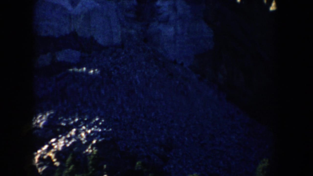 Blue-toned 8mm view near Mount Rushmore, with a dim rocky slope and trees visible in heavy shadow.