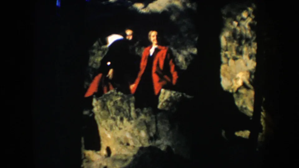 1953 South Dakota home-movie frame showing two adult visitors posing on rocks near trees and stone.