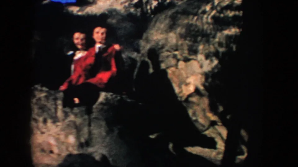 Two adults in red and dark clothing sit or crouch on a rocky ledge in 1953 South Dakota home-movie footage.