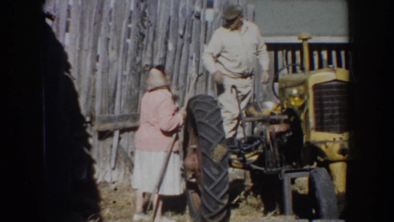 Two adults beside a yellow tractor and rough wooden fence in 1953 South Dakota farm footage.