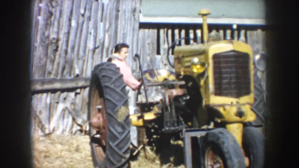 Adult man works near a yellow tractor on a South Dakota farm in 1953, with barn boards and hay-covered ground behind him.