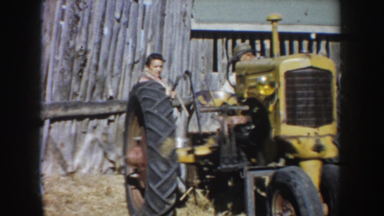 The man stands at the side of the yellow tractor, with the large rear tire and barn wall prominent in the frame.