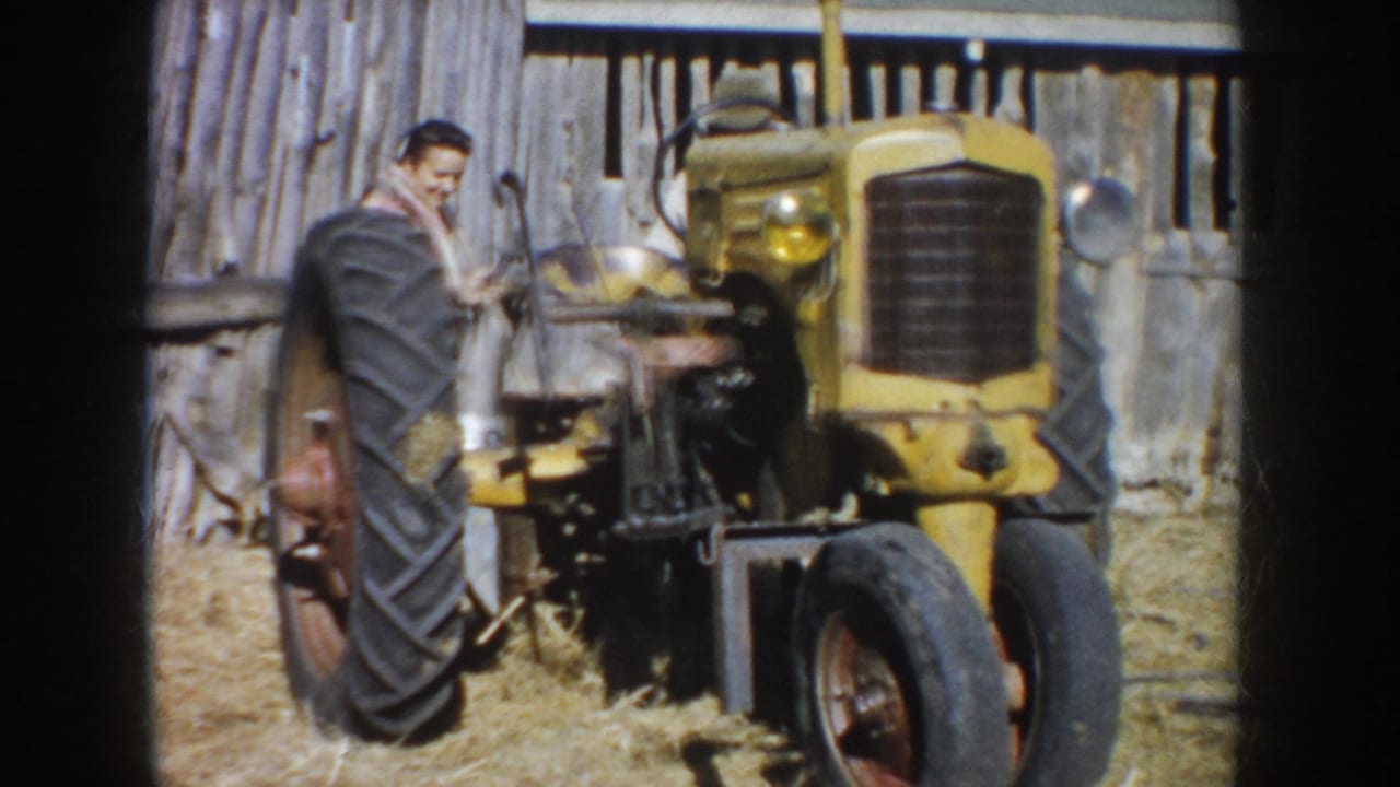 A yellow farm tractor fills the frame, with the man partly visible behind the large rear tire near the barn.