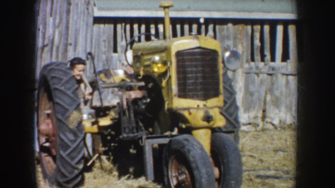 The man leans close to the side of the yellow tractor, with its front grille, wheels, and weathered barn backdrop visible.