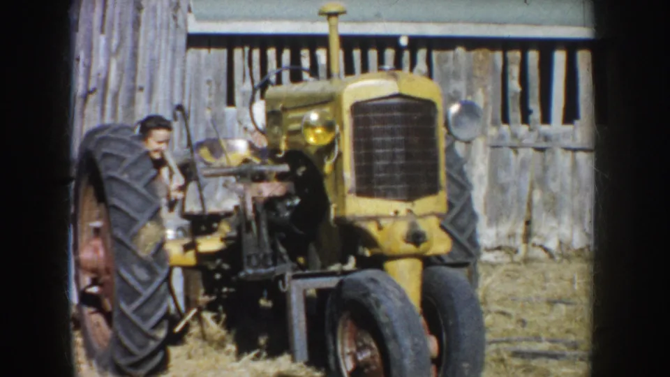 The man leans close to the side of the yellow tractor, with its front grille, wheels, and weathered barn backdrop visible.