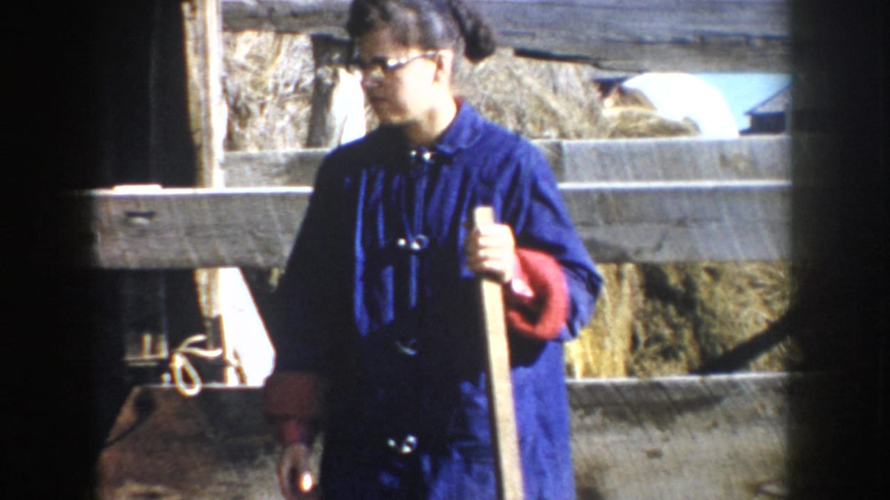 1953 South Dakota 8mm frame showing a woman in blue work clothing holding a long-handled tool inside or beside a barn enclosure.