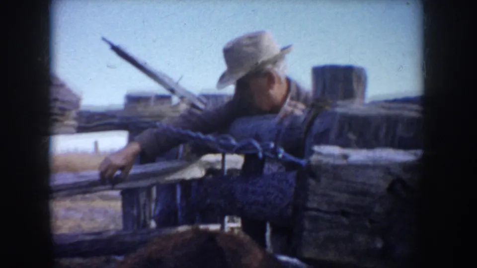 A farmer in work clothes and a wide-brimmed hat leans over a wooden fence near cattle in South Dakota farm footage.