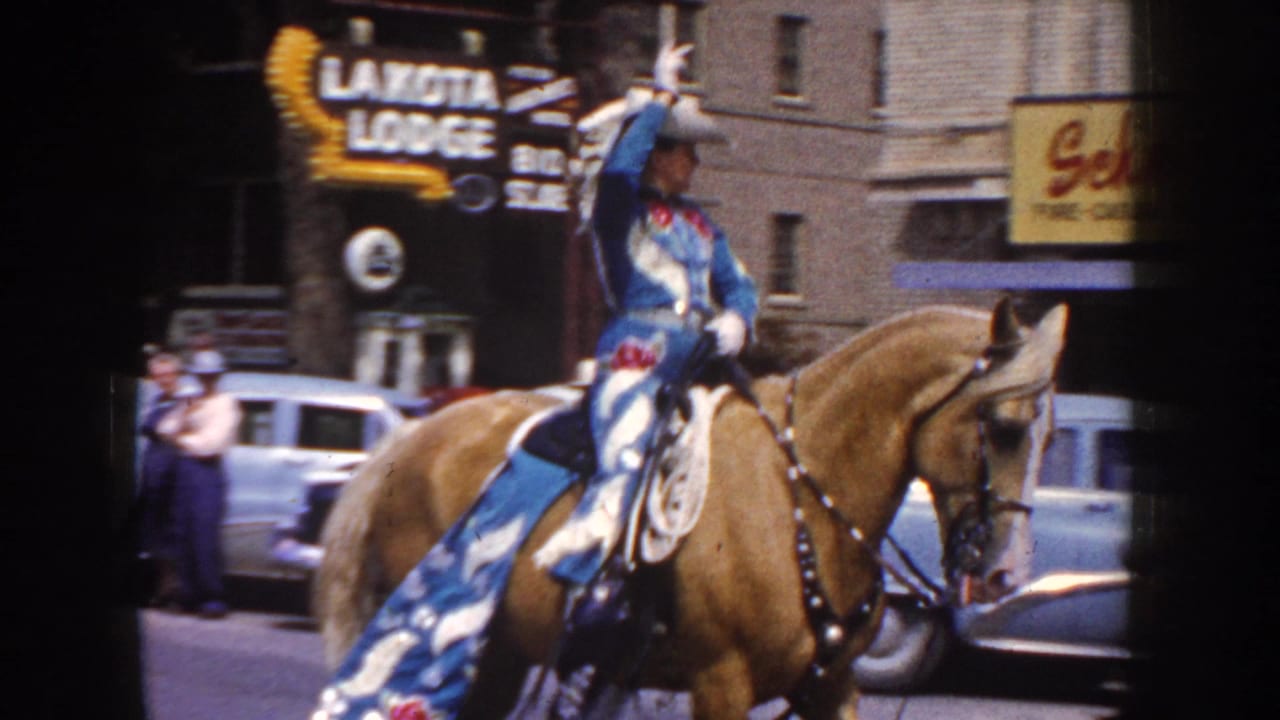 1954 South Dakota parade frame with a cowgirl in blue and white riding a light brown horse past storefronts and parked cars.