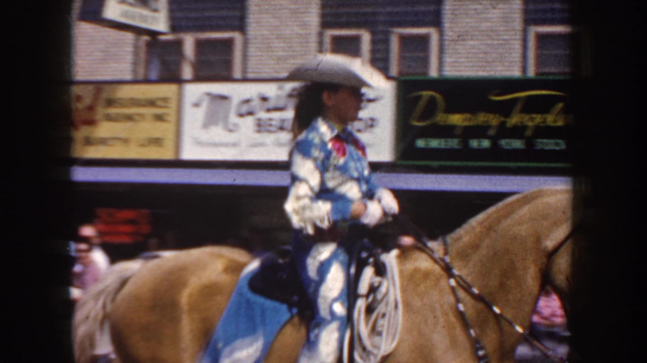 Mounted cowgirl rides past storefront signs and parked cars during the parade.