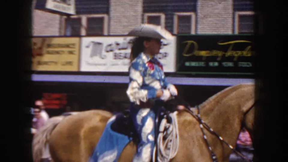 Cowgirl in blue and white costume rides a light brown horse along a South Dakota town street in 1954 parade footage.
