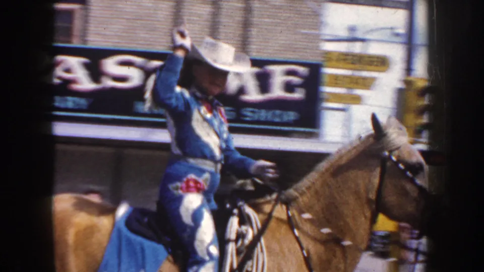A blue-costumed cowgirl raises her white hat from horseback in 1954 South Dakota parade footage with signs behind her.