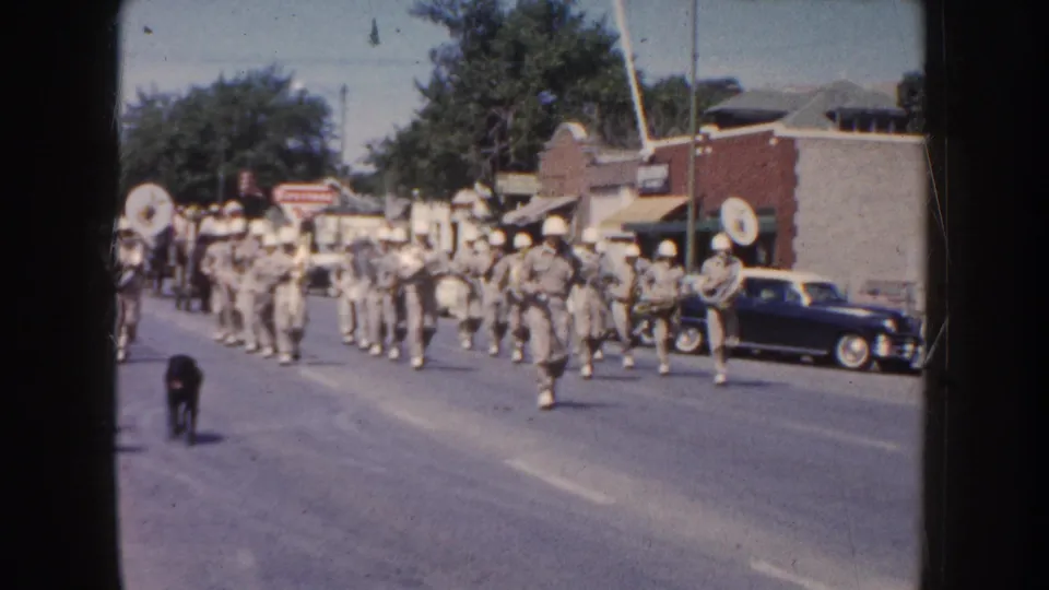 A uniformed marching band follows the parade route on a South Dakota street, with 1950s cars and buildings visible.
