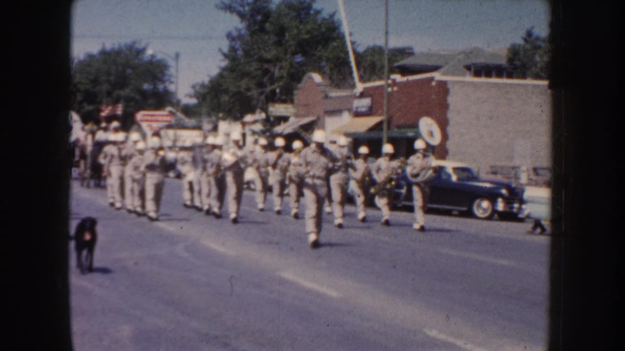 1954 South Dakota parade footage showing a marching band approaching on a main street, with spectators, parked cars, storefronts, and a dog in front.