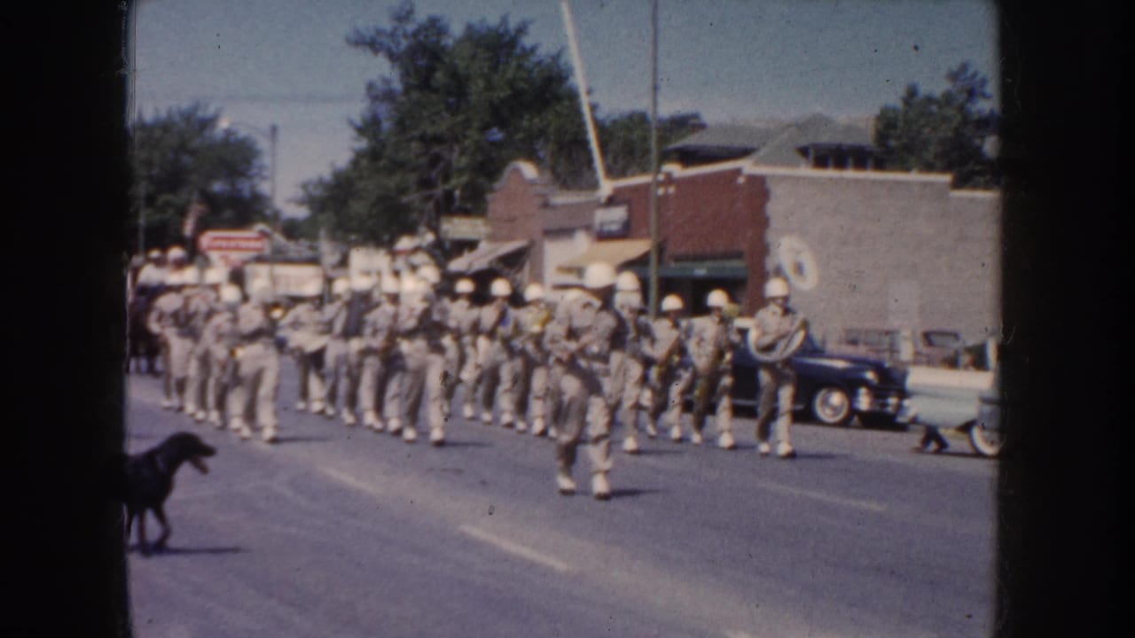 South Dakota 1954 parade view with uniformed band members, brass instruments, a dog in the roadway, parked vintage cars, and people watching from the sidewalk.