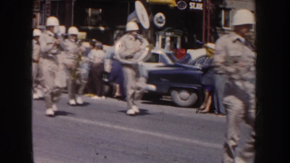 Close home-movie view of a 1954 South Dakota marching band passing spectators, with white helmets, pale uniforms, a tuba, saxophones, and mid-century cars at the curb.