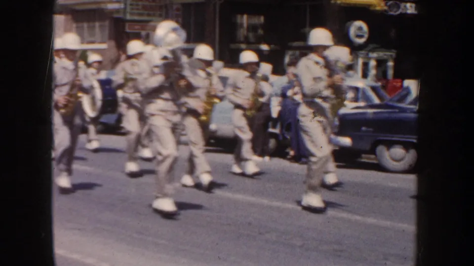 1954 South Dakota street parade frame with uniformed musicians moving past a parked station wagon, a dark sedan, storefronts, and spectators watching from the curb.