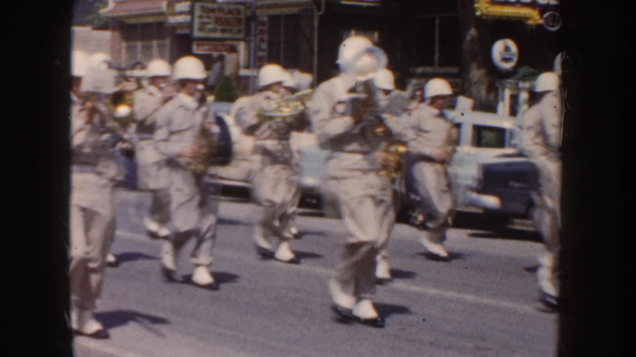 1954 South Dakota home-movie frame of helmeted marching band members playing brass instruments in a street parade.