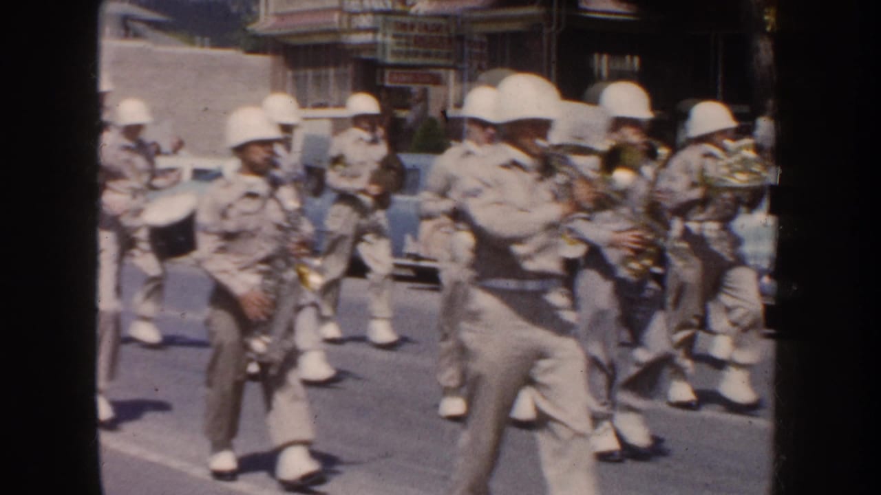 Helmeted musicians march in formation while playing brass and woodwind instruments on a town street.