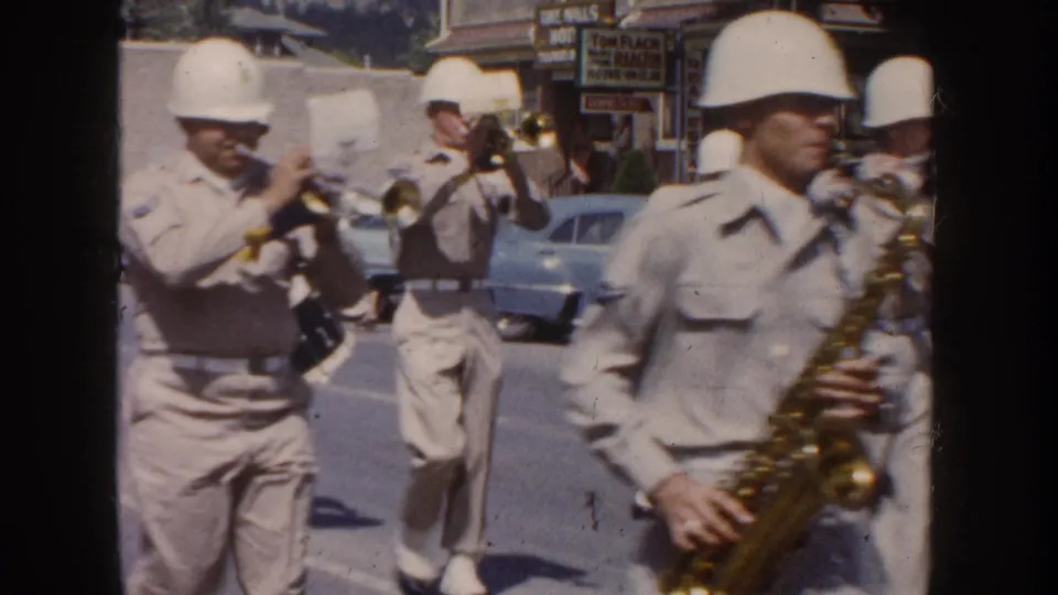 1954 South Dakota parade frame with helmeted band members playing trumpet and saxophone near storefront signs.