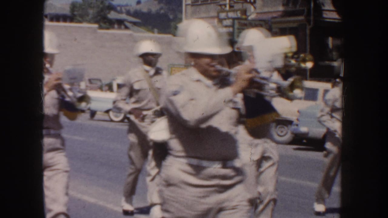 1954 South Dakota home-movie frame showing light-uniformed marching band members with brass instruments on a parade route.
