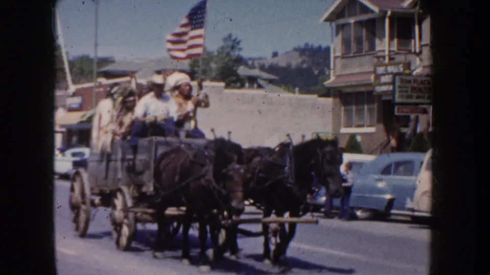 Horse-drawn wagon with seated parade participants moves down a 1954 South Dakota street lined with classic cars.