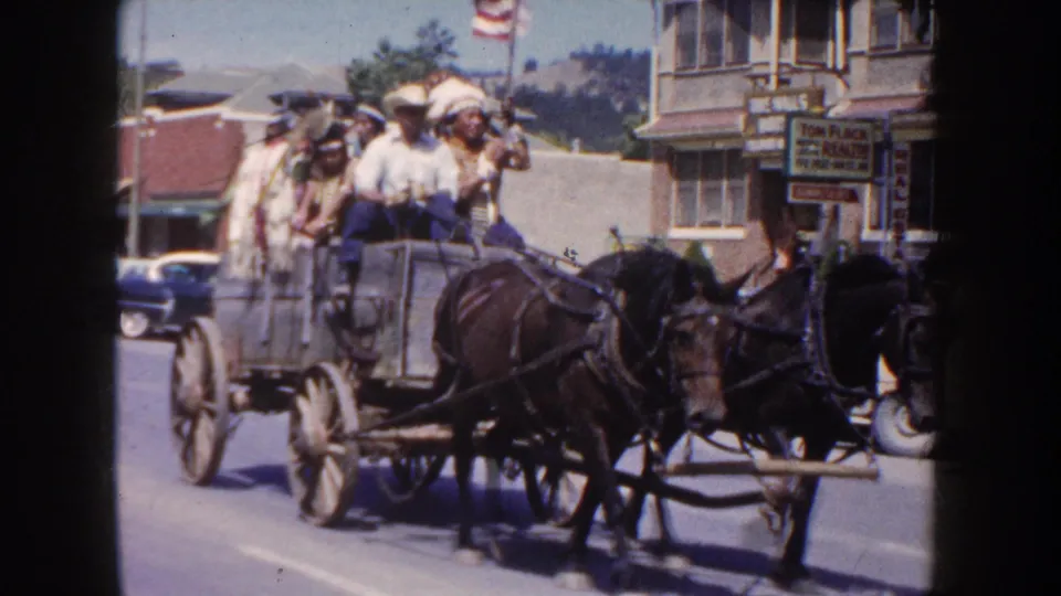 1954 South Dakota parade frame showing a horse-drawn wagon with riders, storefront signs, cars, and an American flag nearby.