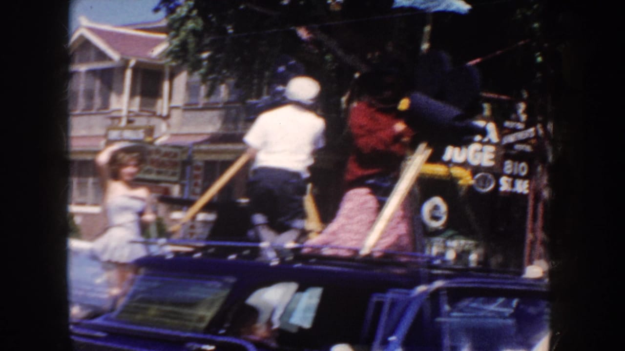 1954 South Dakota parade frame showing a woman waving beside a dark classic car with riders, storefronts, and street signage in view.