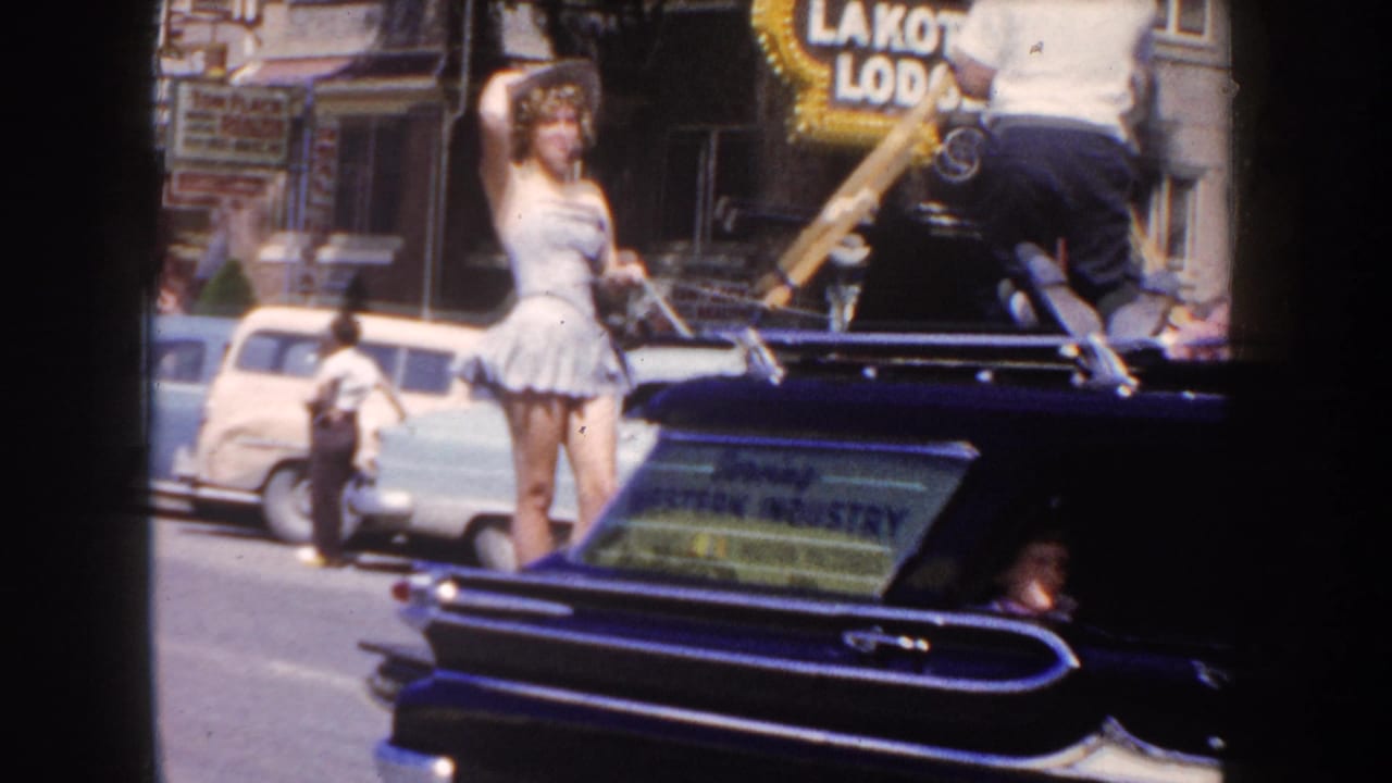 1954 South Dakota parade view with a woman waving from a dark convertible, parked cars, storefronts, and a Lakota Lodge sign visible.