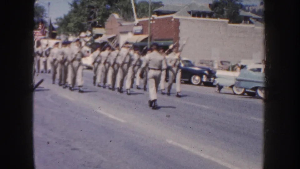 1954 South Dakota street parade frame showing light-uniformed marchers in formation, parked mid-century cars, buildings, and an American flag.