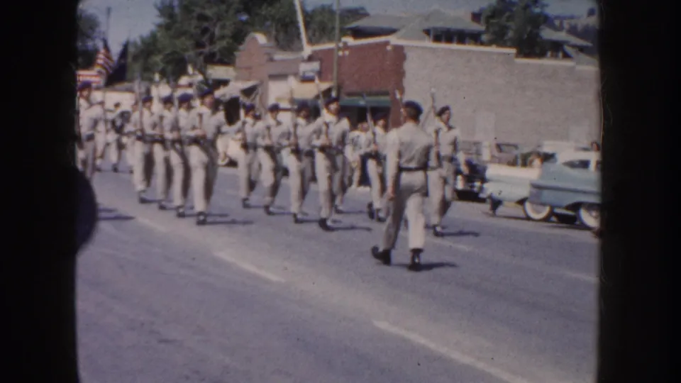 1954 South Dakota parade view of a marching band in light uniforms arranged across the street, with parked cars and low buildings behind them.