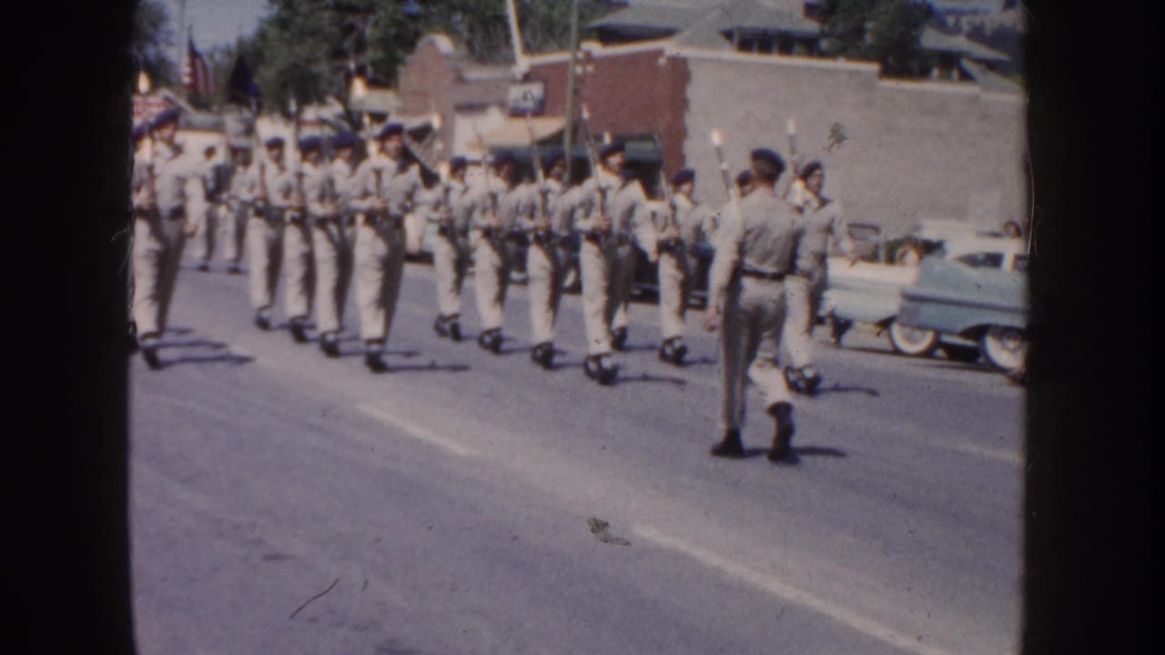 1954 South Dakota home-movie frame showing uniformed young men with rifles forming ranks during a town parade.