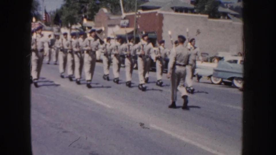 1954 South Dakota home-movie frame showing uniformed young men with rifles forming ranks during a town parade.